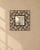 A wrought iron scroll wall mirror, small and square inside a wide frame of black iron scrollwork. Vines and curls repeat...