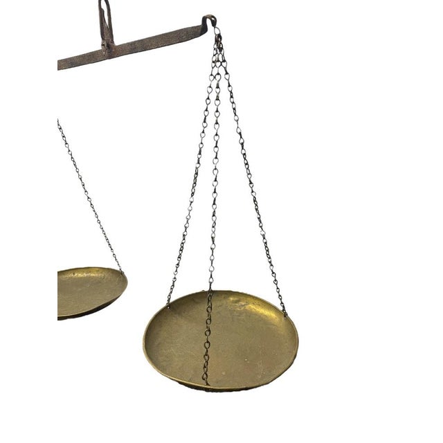 An antique hanging balance scale featuring dual brass pans suspended by chains from a central iron beam. Hanging balance...