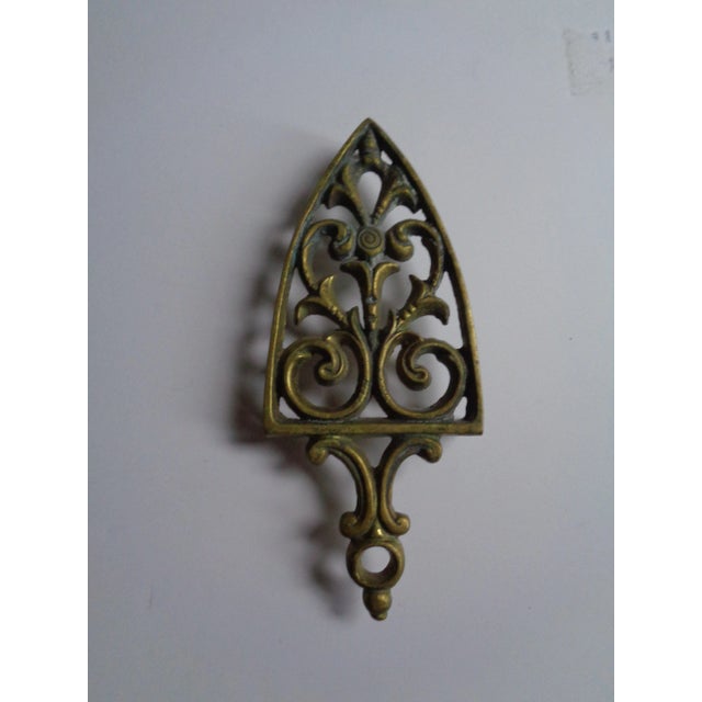 Pretty mid-century trivet. Shield-shaped. Made of brass. With feet and hanging hole at top.