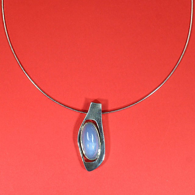 This handcrafted sterling silver pendant showcases a striking long oval blue agate cabochon, beautifully set in a sleek,...