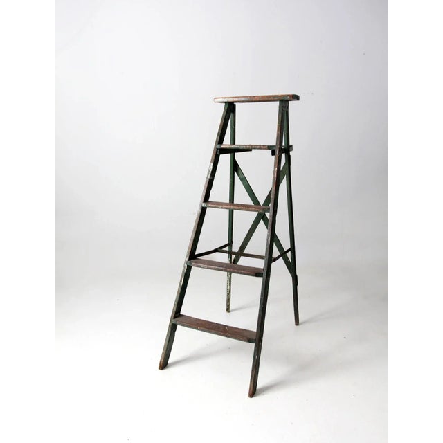 This is a vintage green wooden ladder. The painted a-frame ladder features four rung steps, a back shelf, and crossing...
