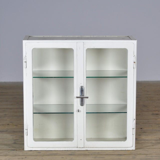 Metal display cabinet dating from circa 1930, featuring a white lacquered finish, two glass doors, and two glass shelves....