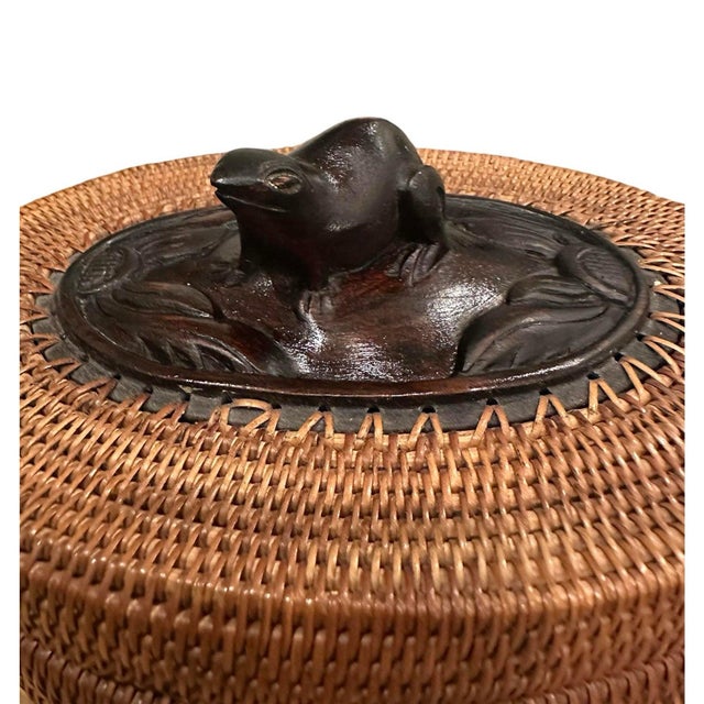 A vintage, Indonesian wicker basket box. Features a handwoven, oval form lidded box adorned with a carved wood frog...