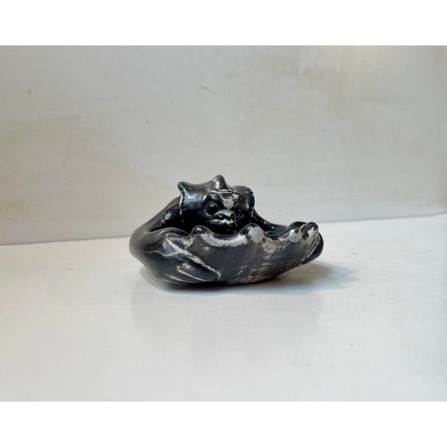 A small sculpted ceramic dish that depicts a devil or demon emerging from a clam shell. Designed and made by the danish...