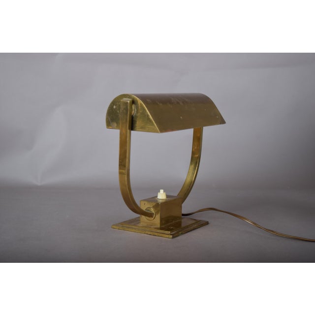 Compact brass desk lamp attributed to the 1930s. A broad U-shaped frame rises from a stepped rectangular base to support a...