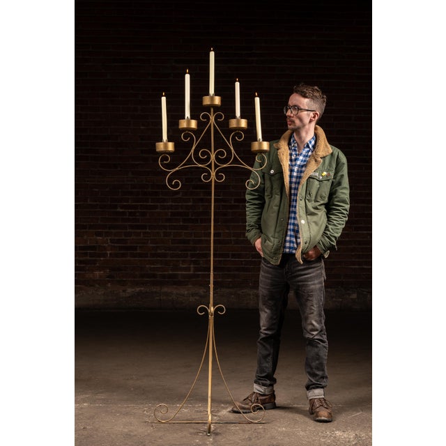 Freestanding wrought iron candelabra stand with a tall, slender central shaft rising from a tripod base formed by scrolled...