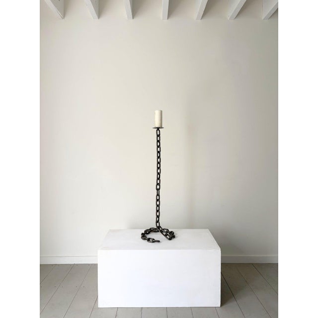 A vintage metal candlestick holder, blacksmith made in the form of a chain. The chain links coil around on the floor to...