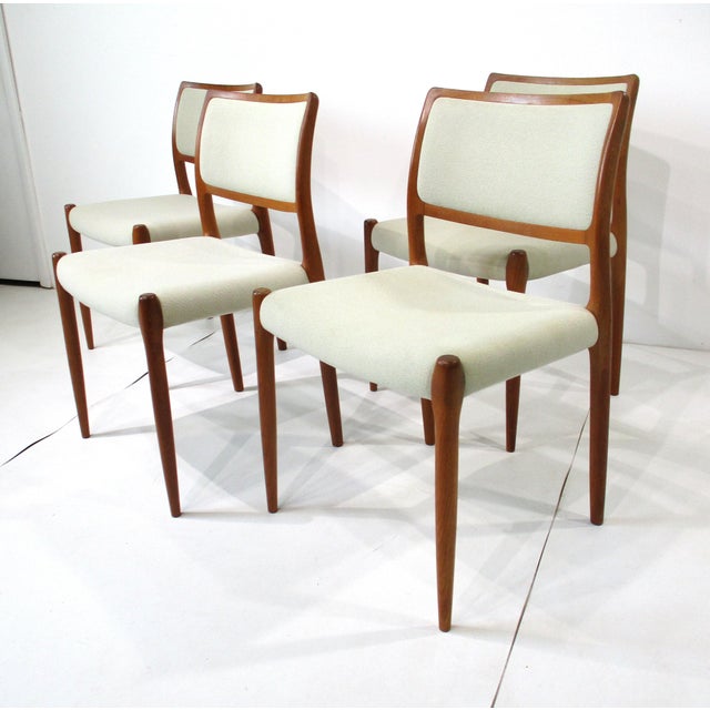 A set of four sculptural teakwood framed dining chairs with woven oatmeal toned fabric back and bottom cushions. An...