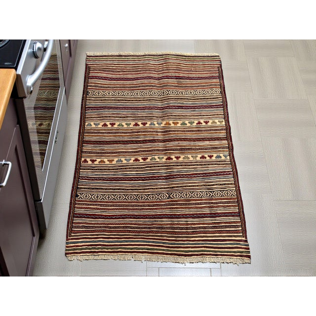 This vintage Afghan Sumak kilim hand‑woven rug displays an all‑over striped field with rows of geometric and abstract...