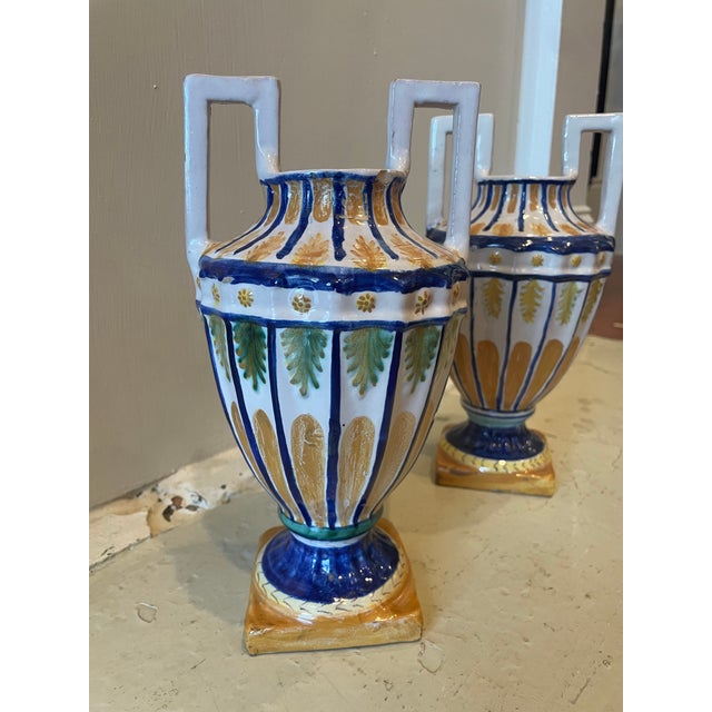 A pair of petite, colorful majolica urns, with distinctive rectangular upright handles and a wide rounded body tapering to...
