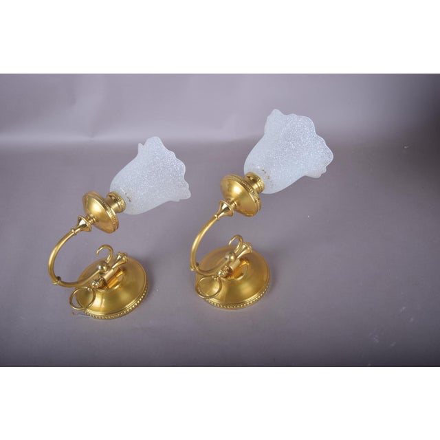Pair of brass wall sconces with beaded round backplates, ring-pull details and single curved arms supporting frosted...