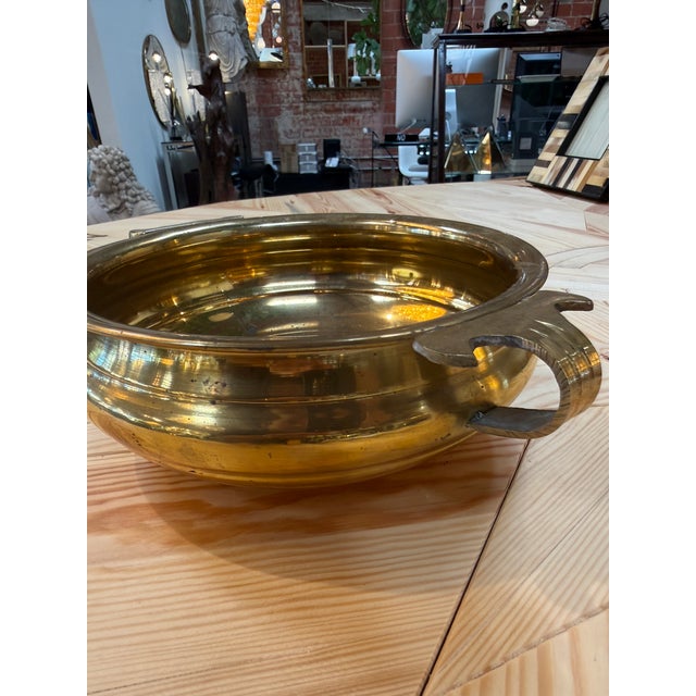An elegant brass bowl featuring a rounded body, a wide rolled rim, and two curved handles accented with stylized leaf...