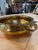 An elegant brass bowl featuring a rounded body, a wide rolled rim, and two curved handles accented with stylized leaf...
