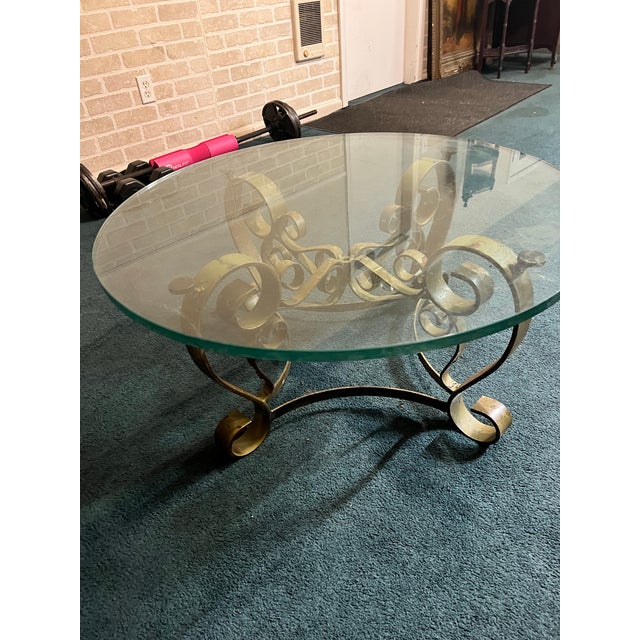 A striking vintage round coffee table featuring a sculptural iron base with decorative scrollwork and a substantial...