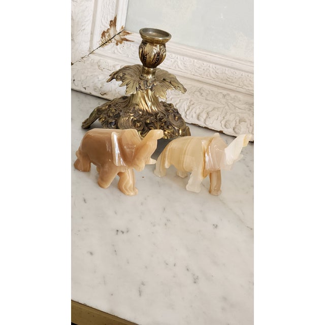 A decorative pair of vintage hand-carved onyx elephant figurines in warm honey and cream banded stone. The raised trunks...