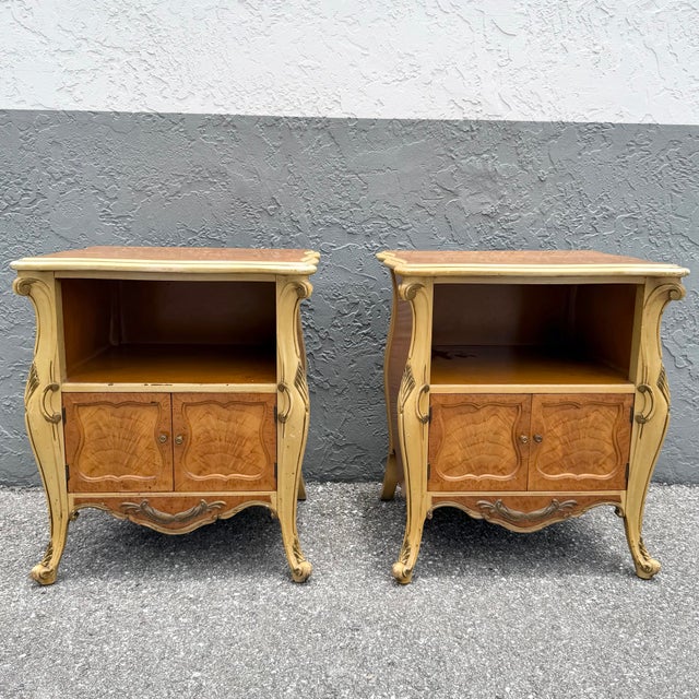 A charming pair of vintage French Provincial style nightstands featuring elegant bombe silhouettes, warm burl wood...