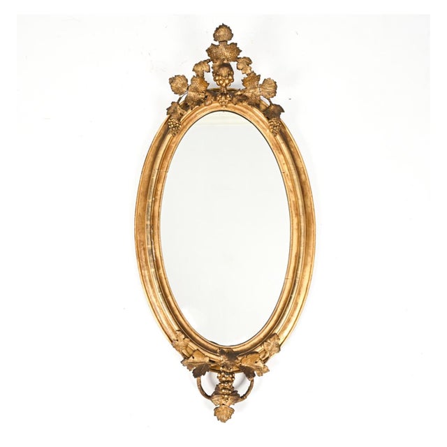 An antique Louis XVI style oval gilt wood wall mirror with a broad, stepped and molded rubbed gold frame with naturalistic...