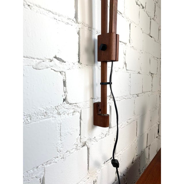 Vintage wall-mounted lamp crafted from teak wood, featuring a distinctive curved arm and a warm yellow fabric lampshade....