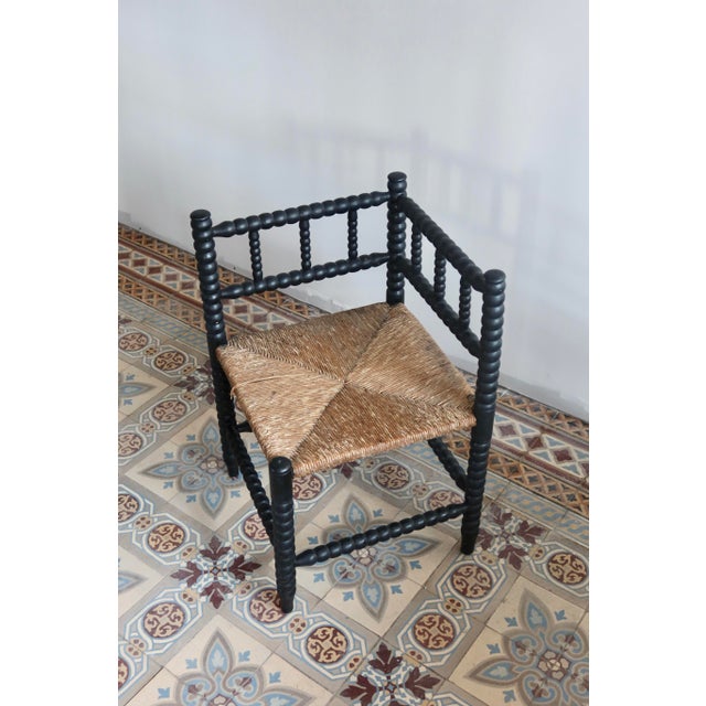 Antique corner chair in turned wood, painted black, with a woven straw seat decorated in a cross pattern. The chair shows...