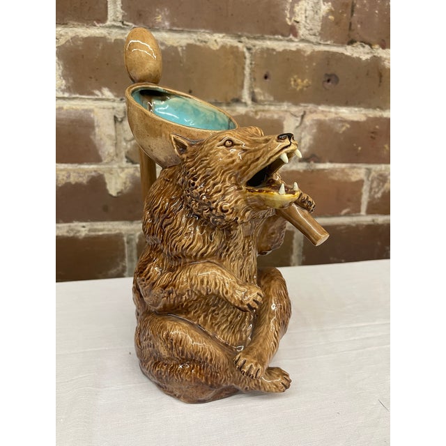 The majolica pitcher is a seated, cross-legged brown bear holding a spoon with a bright blue interior. The handle is...