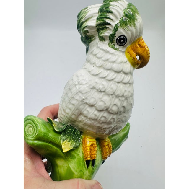 This is a charming porcelain figurine depicting a white parrot perched on a vibrant green tree stump. The parrot features...