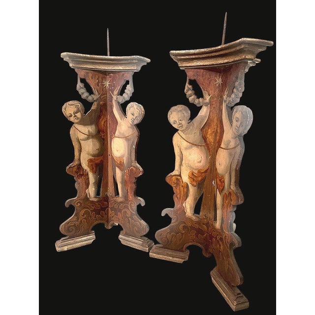 Pair of large palace candle holders in painted wood, featuring shaped representations of triumphant putti holding a...