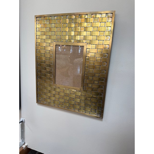 A decorative wall frame with a richly textured brass surround. Rows of rounded, interlocking shapes create a woven effect,...