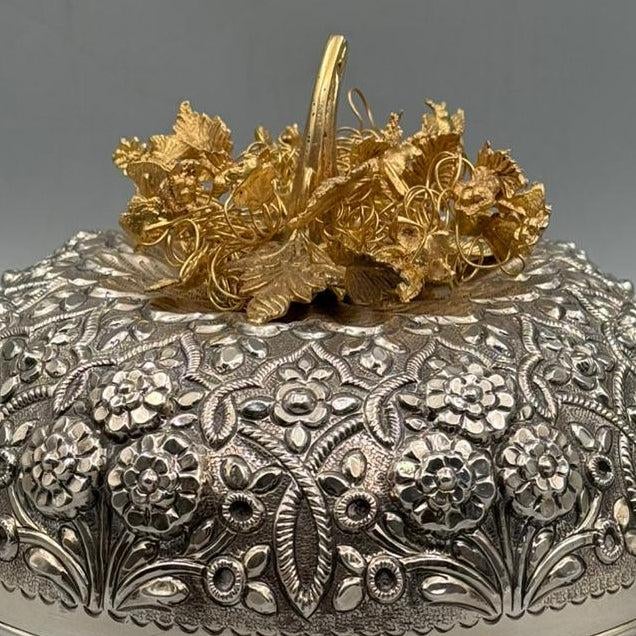 A richly hand-engraved bonbonniere crowned with an intricate gold-plated floral arrangement. The body is covered in dense...