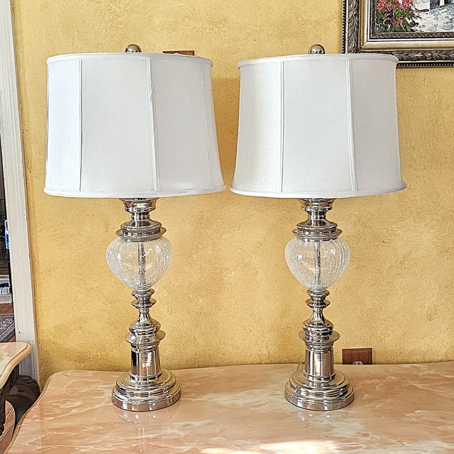 A matching pair of tall table lamps featuring polished chrome bases with crackle glass globe accents and crisp white...