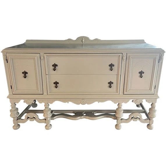 Vintage sideboard featuring a soft warm beige painted finish with carved detailing and turned legs. Two center drawers are...