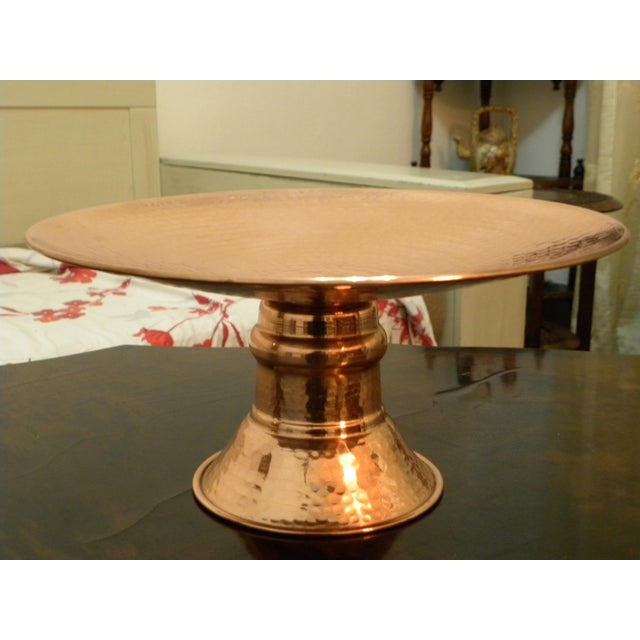 A elegant hand hammered copper pedestal cake stand featuring an authentic, elegant design. Its circular tray rests on a...