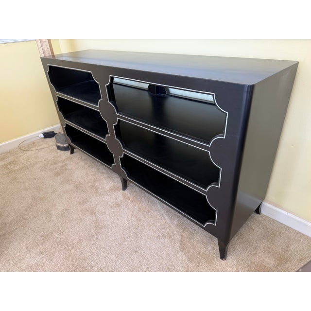 The striking console features three long shelves framed by a striking trellis‑like front, finished in a smooth black...