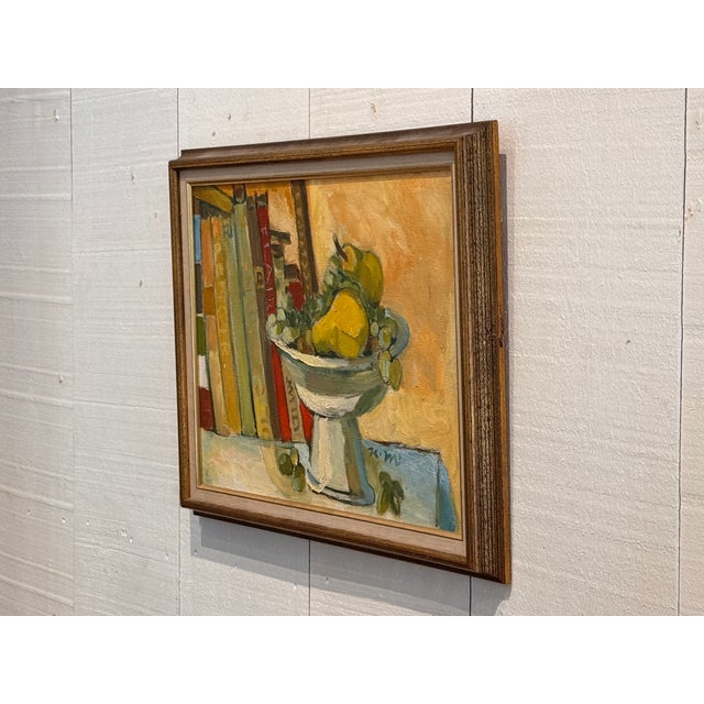 Oil on canvas still life showing a footed bowl of fruit — pears and grapes — set against a row of books, painted in warm...