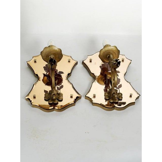Pair of French wall sconces in patinated gold metal and tinted mirrored glass, featuring an elegant stylized floral...