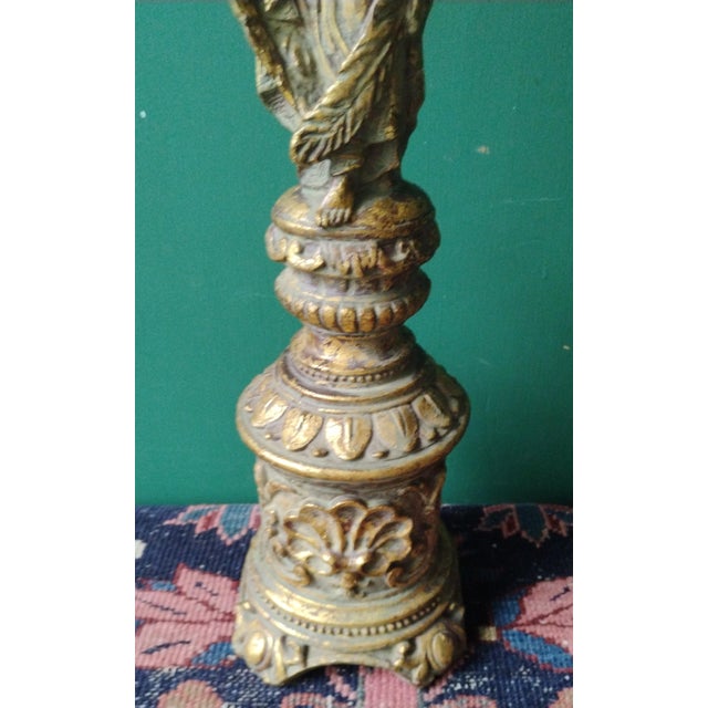 An elaborately carved tall candlestick holder featuring raised angel figures in a prayerful pose. The design showcases...