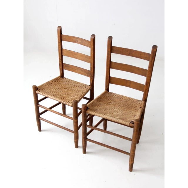This is a pair of antique splint weave seat chairs. The rustic wooden chairs feature ladder backs and splint weave seats...