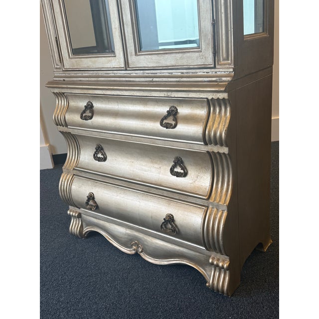 Elegant champagne-finished display cabinet featuring ornate French-inspired detailing and a beautifully curved crest. The...