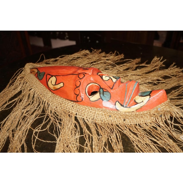 A striking decorative wall mask featuring an elongated, expressive face surrounded by dramatic natural-fiber fringe. The...