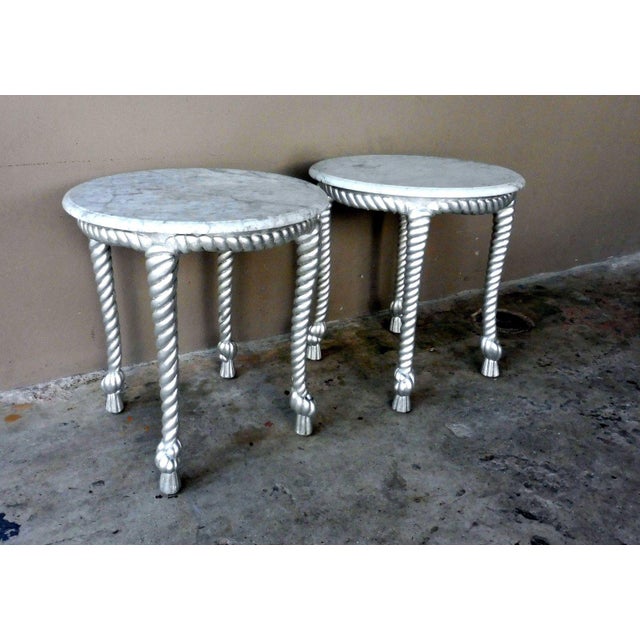 This is a pair of Hollywood regency tables from the 1970s. The pieces feature marble tops and silver leafed, twisted rope...