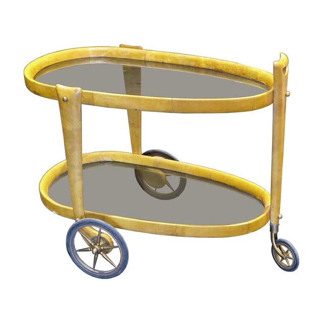 This is a two tier parchment covered wood in oval shaped bar cart by Aldo Tura features three wheels with brass details....