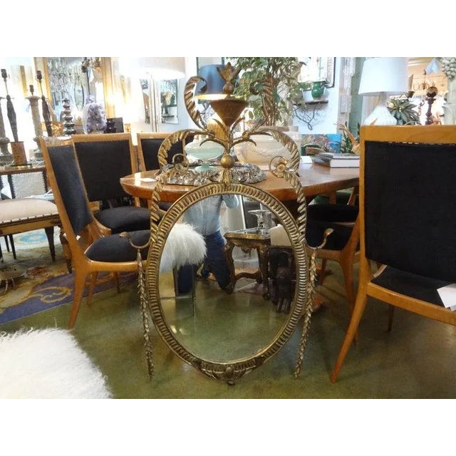 Stunning Italian 1960s Neoclassical style gold gilt metal oval mirror with trophy crest and floral and foliate swags. This...