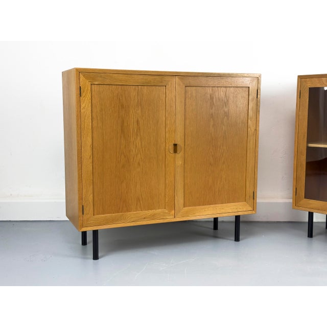 Two compact, two-door oak cabinets, one with glass doors, both with adjustable shelves. Product Information: •...