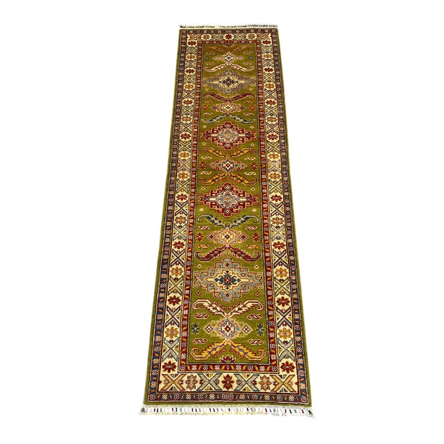 This natural dye wool runner features an olive green field with repeating Kazak-style tribal motifs. A cream border adds...