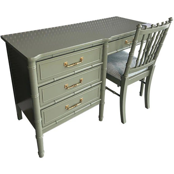 A compact painted desk in a muted green finish with three drawers and simple brass pulls, paired with a matching spindle-...