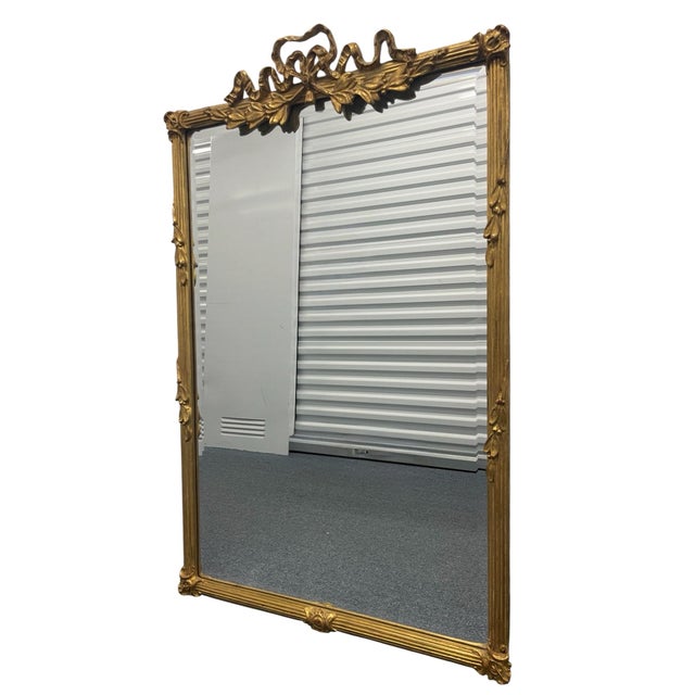 A refined Carvers’ Guild wall mirror featuring an ornate ribbon-and-foliate crest in a warm old gold leaf finish. The...
