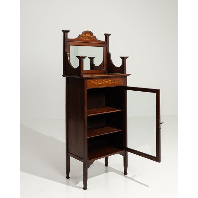 This display cabinet makes its impression through dark mahogany with inlay, glass door, open shelf. The piece dates to...