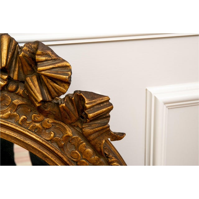 A large oval gilt wood wall mirror with an ornately carved frame topped with a decorative bow-and-ribbon crest and...