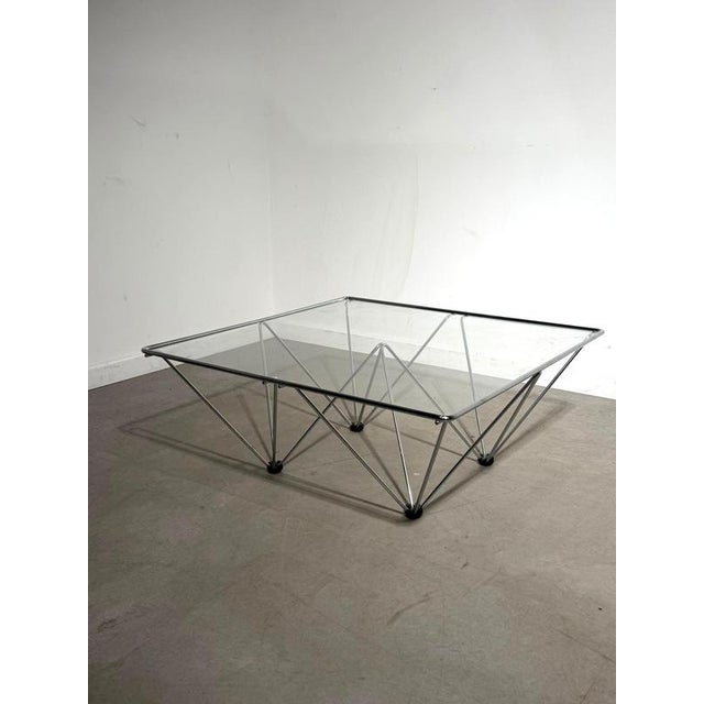 Striking square coffee table featuring a sculptural geometric chrome frame and clear glass top. The open metal...
