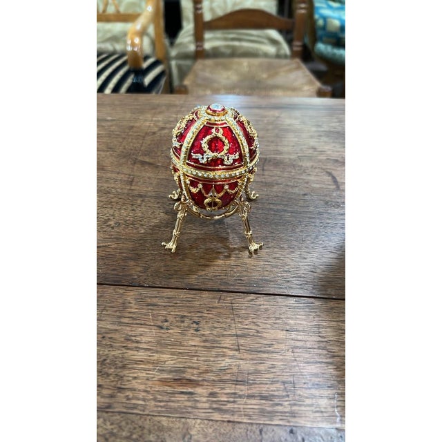 An exquisite Fabergé decorative egg trinket box featuring rich ruby-red enameling, gold-tone filigree, and sparkling...