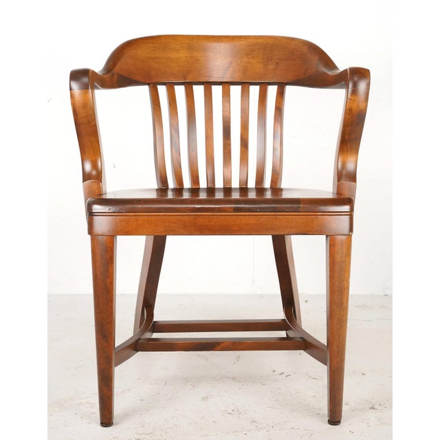 Vintage banker chair made of wood in a dark tone stain. It features a slatted back, curved arms, a contoured seat, and...